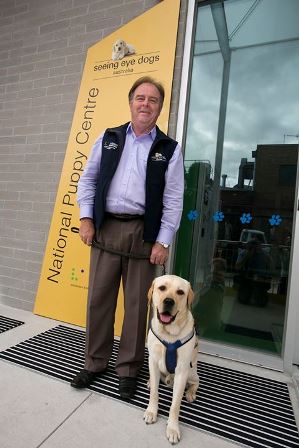 Leigh stands in front of Seeing Eye Dogs puppy centre with a golden Labrador Seeing Eye Dog by his side.