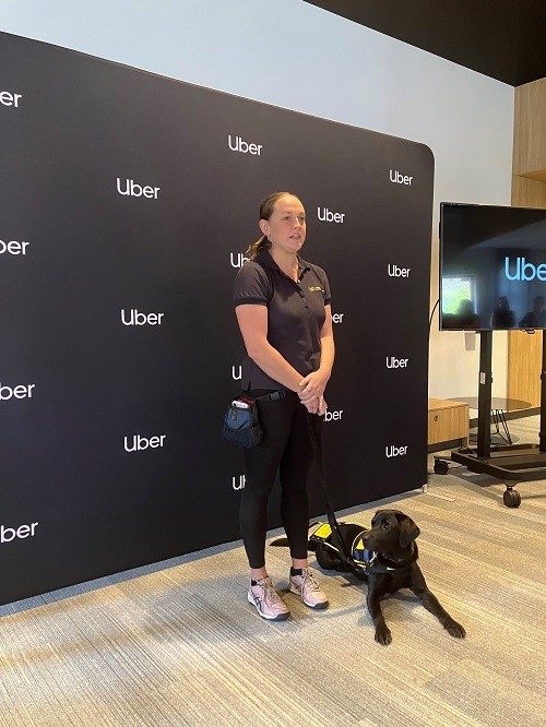 A woman stands at the front of a room with a Seeing Eye Dog in training