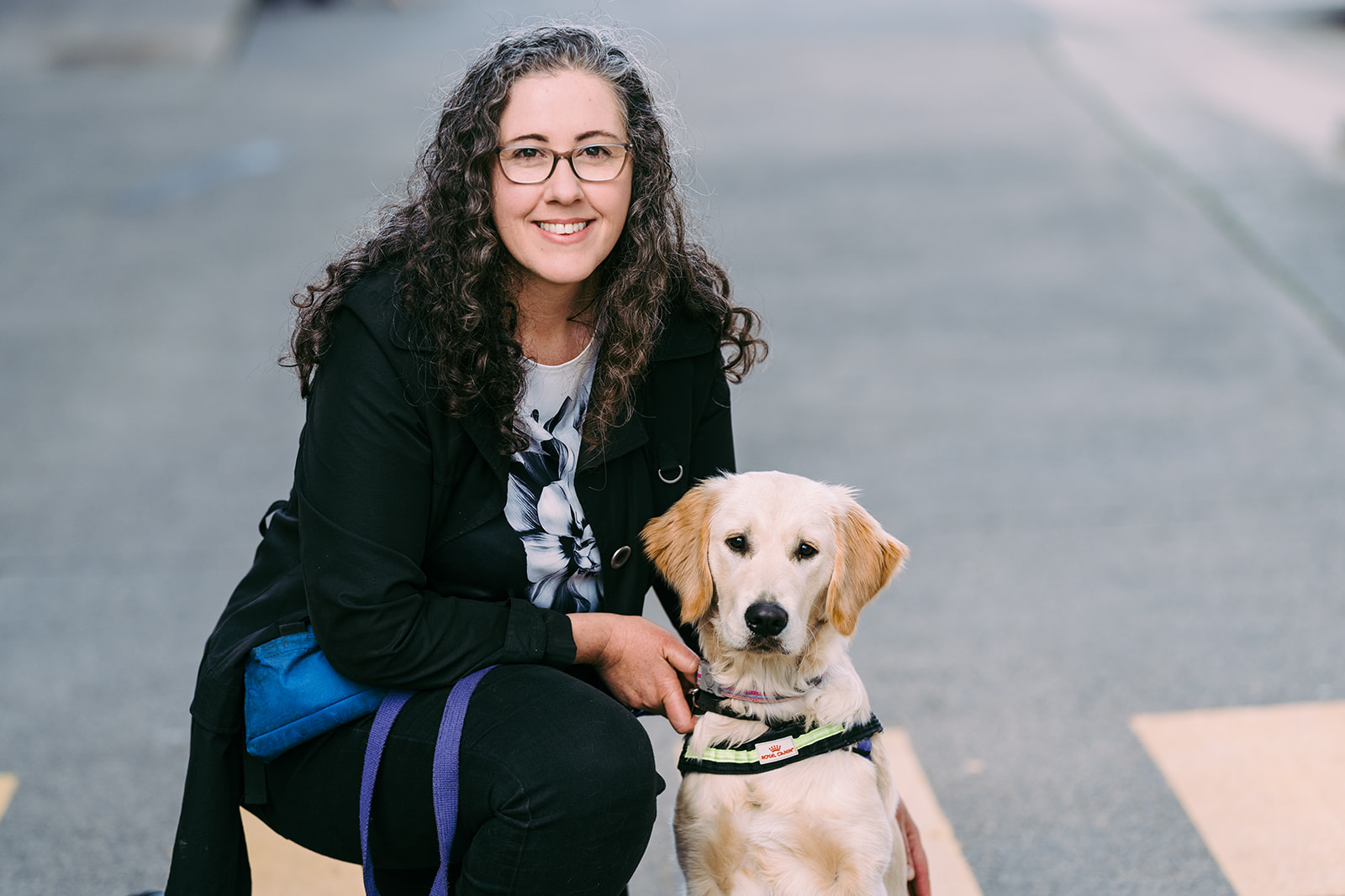 A woman with long dark curly hair and wearing glasses is smiling at the camera. She is crouched beside a Seeing Eye Dogs puppy in harness